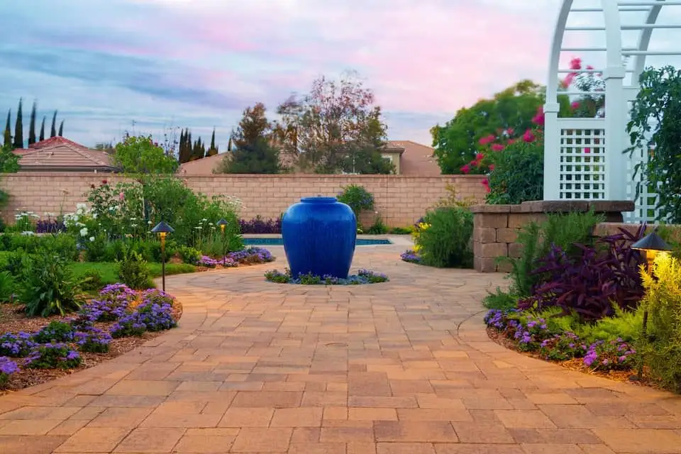 A large, blue, glazed, ceramic pot that has been converted into a fountain sits in the middle of a paved path surrounded by a small, colorful garden.