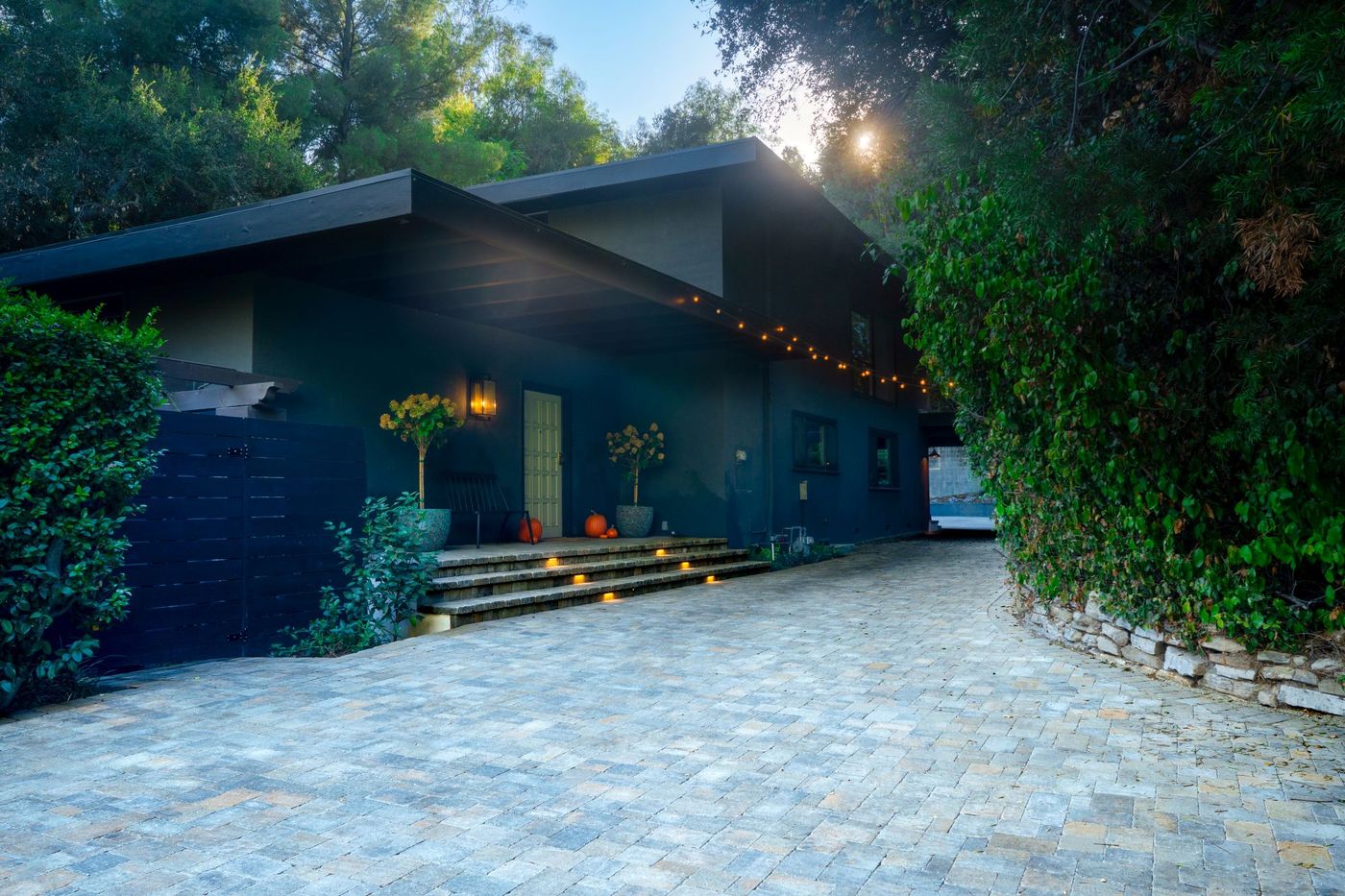 Wide residential paver driveway leading to a modern home with integrated step lighting, string lights, and lush surrounding greenery