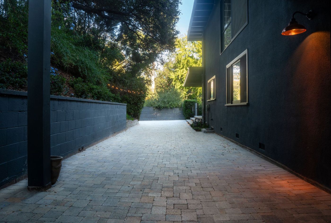 Long paver walkway along a contemporary dark-painted home with retaining wall, exterior lighting, and surrounding trees